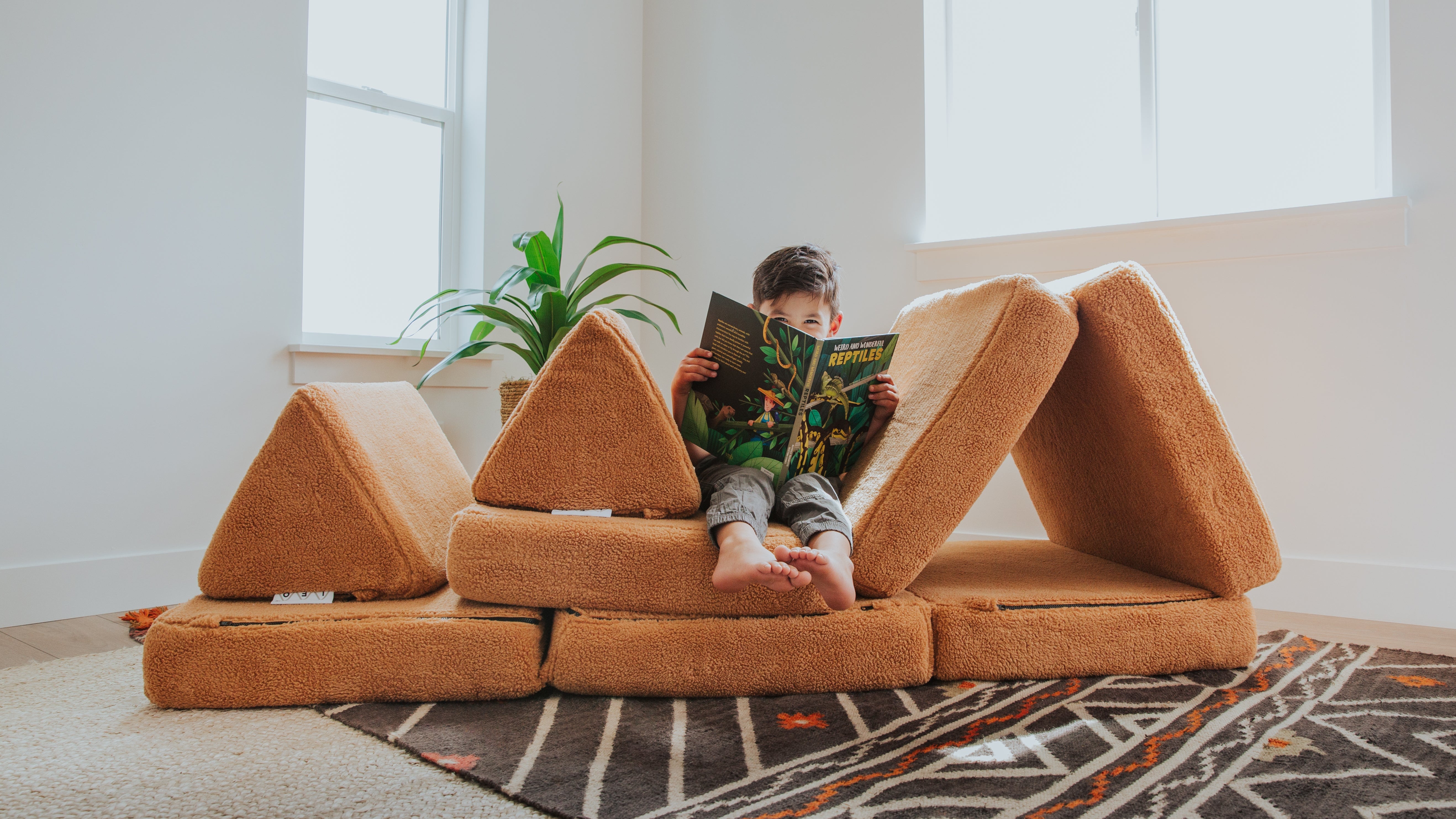 Child reading a book on a brown modular sofa in a bright room with large windows.